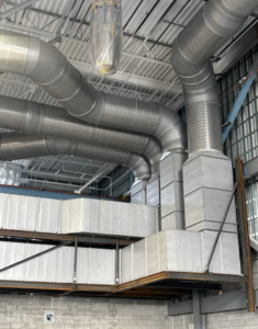 Industrial warehouse interior with large curved silver HVAC ducts running along the ceiling, supported by metal framework above a mezzanine.
